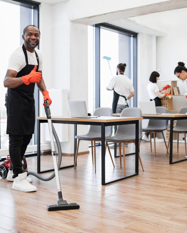 Professional service team of cleaning company. Young adult African American man wearing black apron and red rubber gloves vacuuming floor of modern light office showing thumb up.